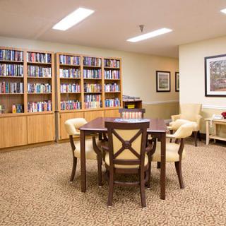 A cozy library room with bookshelves, a table, and chairs arranged on a carpeted floor.