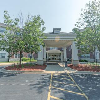 A building entrance with a covered drop-off area, surrounded by trees and landscaping, adjacent to a parking lot.