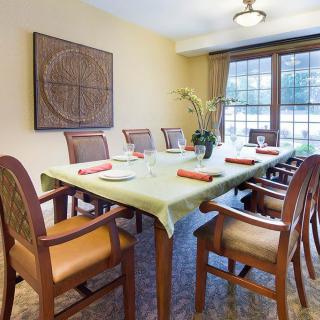 A neatly arranged dining room featuring a long table with chairs, decorated with a green tablecloth and floral centerpiece. Large windows provide natural light, and artwork adorns the wall.