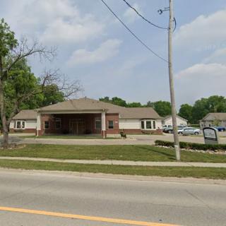 A suburban commercial building with a parking lot, situated along a road lined with trees.