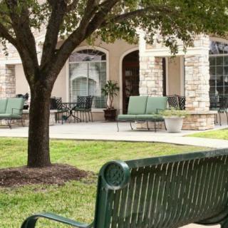 A peaceful outdoor seating area with a green bench under a tree, adjacent to a patio with chairs.
