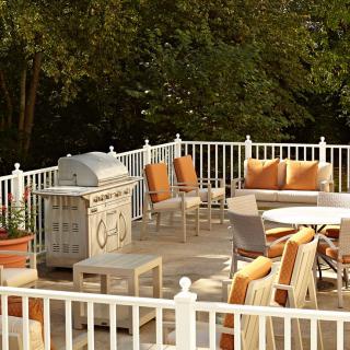 A sunlit outdoor patio with a grill, table, and cushioned chairs surrounded by trees.