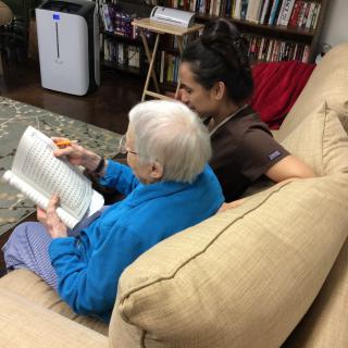 An elderly person reads a book with a caregiver sitting beside them on a couch.