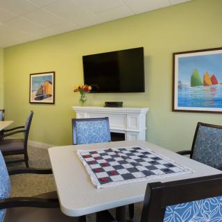 A cozy room with a chess board on the table, surrounded by chairs and a TV mounted on a light green wall adorned with colorful artwork.