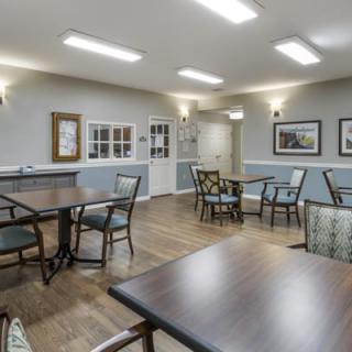 A well-lit dining area with tables and chairs, featuring paintings on the walls and a wood floor.