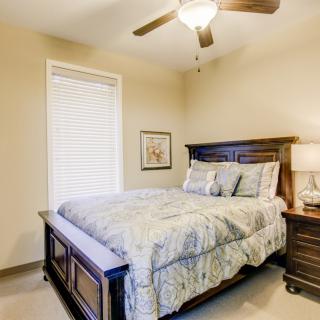 A cozy bedroom with a wooden bed frame, complemented by a matching nightstand and lamp, featuring a large window and ceiling fan.