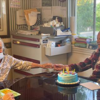 An elderly couple celebrates a birthday with a cake and candles in their cozy dining room.