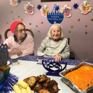 Two elderly women enjoying a Hanukkah celebration with traditional food and festive decorations.