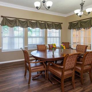 A spacious dining room featuring a large wooden table, surrounded by chairs, with windows adorned with curtains and two chandeliers providing lighting.
