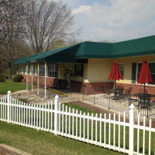 A cozy restaurant with outdoor seating under a green awning, featuring red umbrellas and a white picket fence.