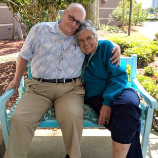 An elderly woman and a man sitting closely on a bench outdoors, sharing an affectionate moment.