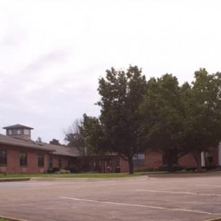 A view of a brick building with a parking lot and trees under a cloudy sky.