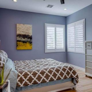 A modern bedroom with a patterned bedspread, abstract artwork on the wall, and white shutters on the windows.