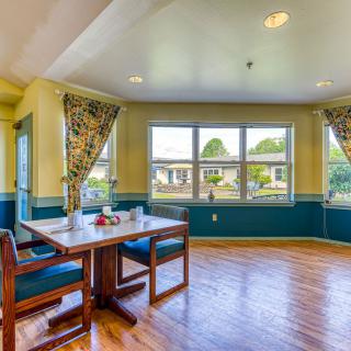 A cozy dining room with a wooden table and chairs by a large window, showcasing bright daylight and decorative curtains.