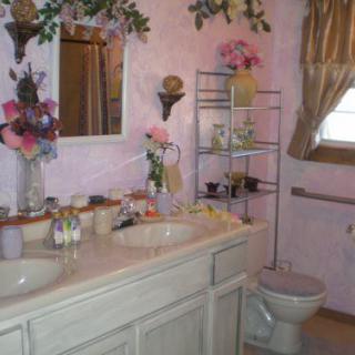 A cozy bathroom with pink walls and floral decorations, featuring a double sink and wooden shelving.