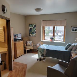 A well-lit and inviting hospital patient room, featuring a bed, seating area, and natural light from the window.