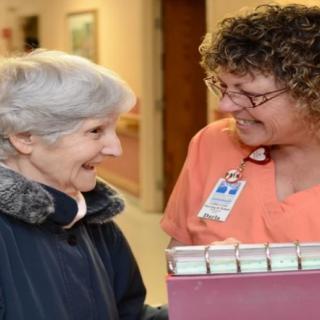 A nurse smiling and interacting with an elderly woman in a healthcare setting.