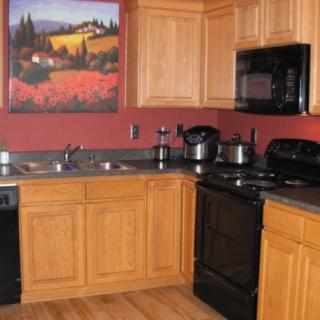 A modern kitchen with wooden cabinets, black appliances, red walls, and a colorful painting above the counter.