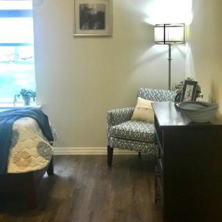 A cozy living room corner with a patterned armchair, standing lamp, and wooden dresser.