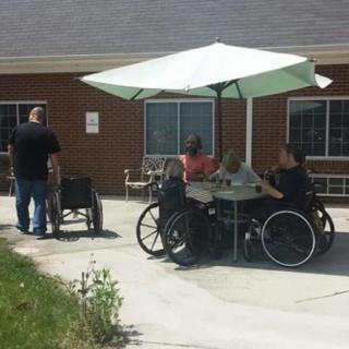 A group of people in wheelchairs sitting at a table with an umbrella outside a building.