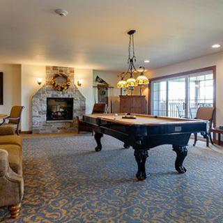 A spacious living room featuring a billiards table, cozy fireplace, elegant chandelier, and large windows offering ample natural light.