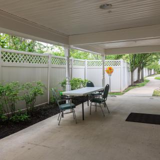 A covered patio space with a table and chairs, surrounded by greenery and a white fence.