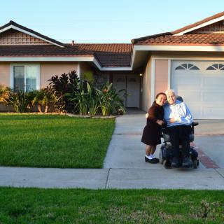 Two people in front of a house, one in a wheelchair, enjoying the sunset.