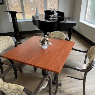 A cozy room with a wooden table surrounded by chairs, featuring a piano in the background and large windows letting in natural light.