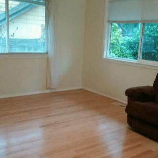 An empty living room with hardwood floors, a single armchair, and large windows letting in natural light.