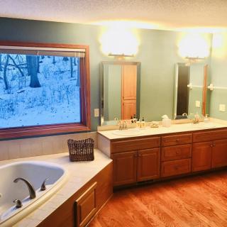 A spacious bathroom featuring a large bathtub, double sinks, and wooden cabinetry with a snowy outdoor view.