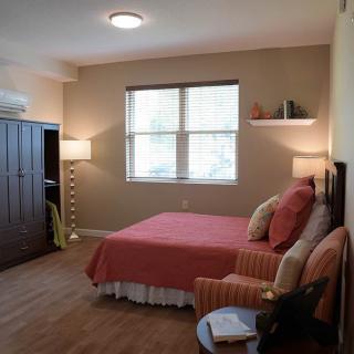 A cozy bedroom with a neatly made bed, armchair, and ambient lighting, featuring warm tones and minimalistic decor.