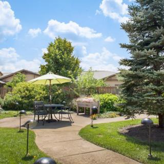 A tranquil backyard garden with a patio set under an umbrella, surrounded by lush greenery and a clear blue sky.