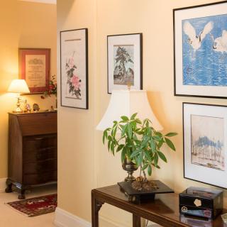 A warmly lit hallway featuring framed artwork, a small table with a lamp and plant, and a cozy corner with furniture in the background.