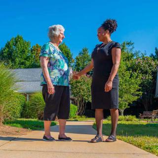 An elderly woman and a younger woman shake hands outdoors on a sunny day.