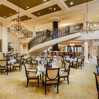 An elegant restaurant interior featuring chandeliers, a grand staircase, and neatly arranged tables and chairs.