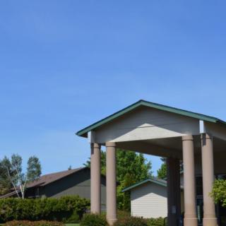 A building with tall columns and a prominent roof structure, set against a clear blue sky with surrounding greenery.