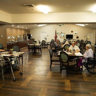 A dining hall with seniors seated at tables, some in wheelchairs, enjoying a meal in a community setting.