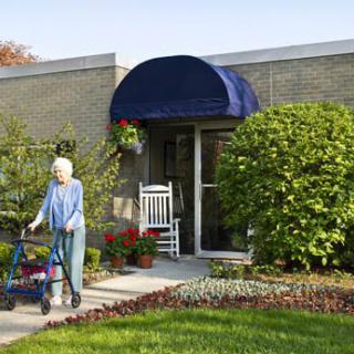 An elderly person using a walker outside a house with a garden and an awning.