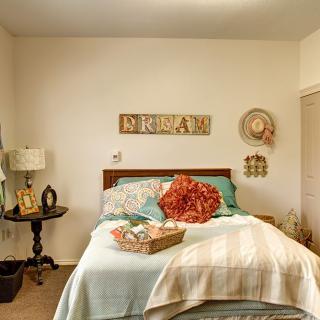 A cozy bedroom with a neatly made bed, featuring floral pillows and a bouquet. The room is complemented by wall art and decorative elements.
