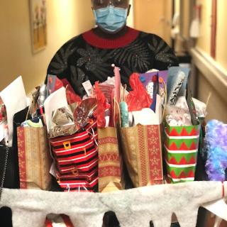A masked individual stands in a hallway, proudly presenting a cart filled with beautifully wrapped holiday gifts.