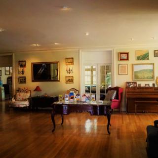 A cozy living room with wooden floors, featuring a piano, a table, and various artworks on the walls.