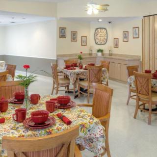A cozy dining room with tables set for a meal, featuring red tableware and floral decorations.