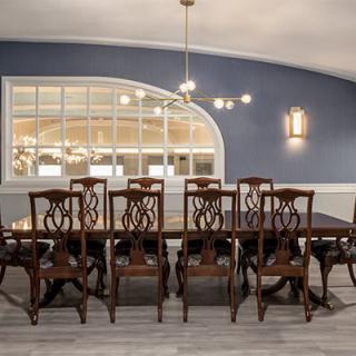 A beautifully decorated dining room with a large wooden table and matching chairs, featuring a blue accent wall and an arched window reflecting light from the chandelier.