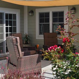 A cozy patio with wicker furniture, surrounded by blooming flowers and shaded by a sun canopy.