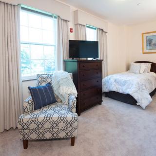 A cozy bedroom with a bed, chair, dresser, and a television near the window.