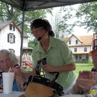 A group of people gathered under a tent during an outdoor event, with a woman using a microphone.