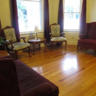 A cozy living room with hardwood floors, featuring a sofa, chairs, a small table, and large windows with curtains.