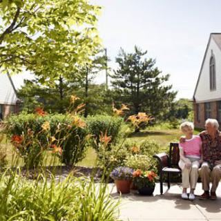 An elderly couple sits on a bench in a beautiful garden, surrounded by vibrant flowers and lush greenery.