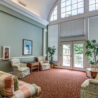 A cozy living room with large windows allowing natural light, furnished with comfortable seating and decorated with plants.