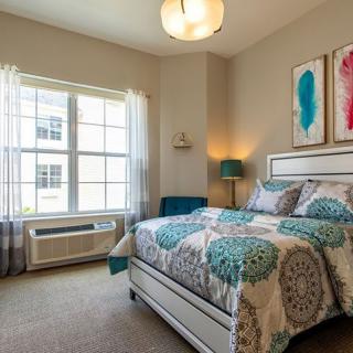 A cozy bedroom with a patterned bedspread, large window, and colorful artwork on the walls.